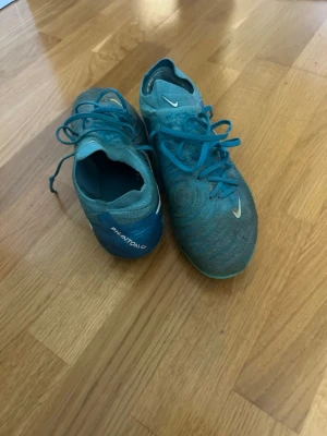 Nike Phantom GT blå fotbollsskor - Säljer ett par Nike Phantom GT fotbollsskor elite i blått med vita detaljer. Skorna har snörning, låg profil och mönstrad ovandel för bättre bollkontroll. Yttersulan har fasta dobbar för spel på konstgräs . Perfekta för dig som vill ha bra grepp och kontroll på planen.