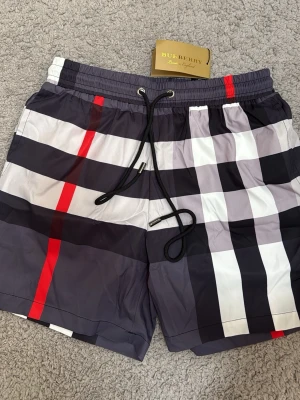 Rutig badshorts från Burberry - Säljer ett par snygga badshorts från Burberry med klassiskt rutigt mönster i svart, vitt, grått och röda detaljer. Shortsen har snörning i midjan och en bakficka. Perfekta för sommaren och strandhäng!