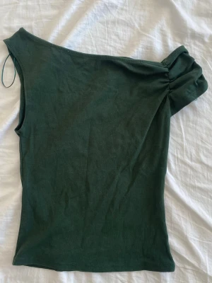 Mörkgrön oneshoulder topp - Snygg mörkgrön oneshoulder topp med rynkad detalj vid axeln. Toppen är ärmlös och har en tight passform som framhäver siluetten. Perfekt för dig som vill sticka ut med en unik och trendig look. Skriv för fler bilder🥰