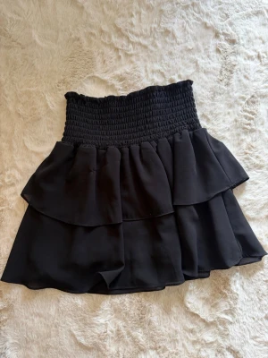 Svart volangkjol från Chelsea - Supersnygg svart kjol från Chelsea med bred smock i midjan och dubbla volanglager. Kjolen har en kort modell och är perfekt för dig som gillar en feminin och trendig stil. Materialet känns lätt och flowy.