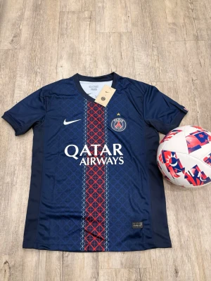 PSG matchtröja blå Nike - Snygg blå PSG matchtröja från Nike med röd och vit detalj i mitten och Qatar Airways-logga på bröstet. Tröjan har korta ärmar, V-ringning och PSG-emblem på vänster sida. Tillverkad i lätt och ventilerande material, perfekt för fotboll eller träning.