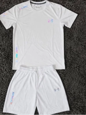 Vitt träningsset från Under Armour - Säljer ett vitt träningsset från Under Armour med t-shirt och shorts. Setet har diskreta loggor i regnbågsfärger och är gjort i ett lätt, snabbtorkande material som passar perfekt för träning. T-shirten har rund hals och shortsen har resår i midjan.
