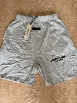 Grå shorts från Essentials Fear of God - Snygga grå shorts från Essentials Fear of God med elastisk midja och logga tryckt på vänster ben. Tillverkade i mjuk bomull för maximal komfort. Perfekta för en avslappnad och trendig stil.
