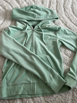 Mintgrön hoodie från Juicy Couture - Snygg mintgrön hoodie från Juicy Couture med dragkedja framtill, huva med dragsko och känguruficka. Mjuk velourkänsla och diskret JC-logga broderad på bröstet. Perfekt för en chill och trendig look.