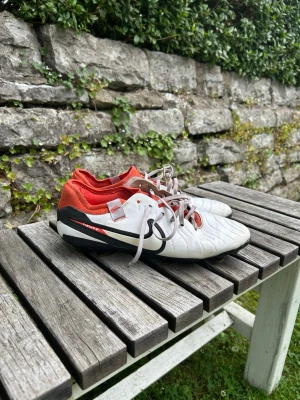 Nike vita fotbollsskor med röd detalj - Säljer ett par vita Nike fotbollsskor med svarta och röda detaljer. Skorna har klassisk snörning, låg profil och är tillverkade i syntetmaterial med en slitstark yttersula. Perfekta för dig som vill ha stilrena och bekväma skor på planen.