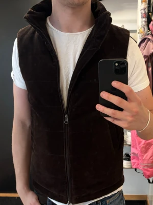Brun Manchesterväst från John Henric - Snygg mörkbrun väst i manchester med hög krage och dragkedja framtill från John Henric. Västen har två fickor på sidorna och en clean, stilren look som passar till många outfits. Pris kan diskuteras!