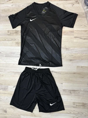 Svart Nike träningsset herr - Snyggt svart träningsset från Nike med t-shirt och shorts. T-shirten har diskreta diagonala ränder och vit Nike-logga på bröstet. Shortsen har resår och dragsko i midjan samt vit logga på benet. Perfekt för fotboll eller gym. Materialet är lätt och andas.