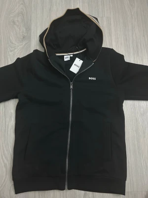 Svart zip hoodie från BOSS - Stilren svart hoodie från BOSS med dragkedja framtill och fickor på sidorna. Huva med beige kantdetalj och diskret BOSS-logga på bröstet. Perfekt för en avslappnad look och enkel att matcha med andra plagg.