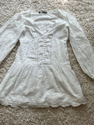 Vit kort klänning med spetsdetaljer - Superfin vit kort klänning med v-ringning, långa ärmar och broderade spetsdetaljer. Klänningen har knappar framtill och en lätt utsvängd kjol med vågig kant. Perfekt för sommarens alla tillfällen.