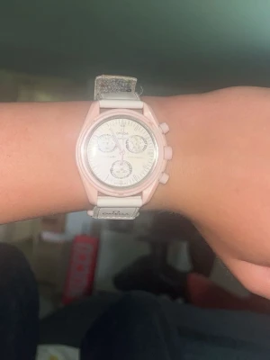 Omega klocka i rosa och vitt - Snygg Omega klocka med rosa boett och vit urtavla. Klockan har flera små urtavlor och ett vitt armband med rosa detaljer. Perfekt accessoar för dig som vill ha något unikt och stilrent på handleden. Helt ok skick behövs fixas lite såsom att byta batteri osv