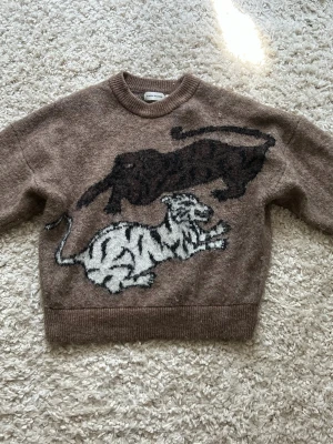 Brun stickad mohairtröja med tigrar - Cool brun stickad mohairtröja från Carin Wester med två stora tigermotiv framtill, en svart och en vit. Tröjan har rund halsringning och ribbade muddar. Materialet är mjukt och ger en avslappnad vibe, perfekt för dig som gillar unika plagg med djurmotiv.