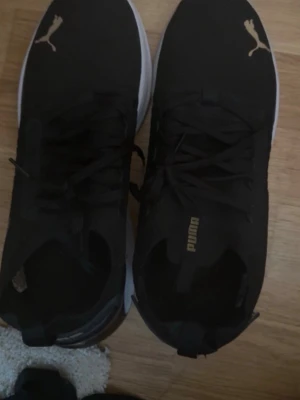 Svart Puma sneaker med vit sula - Snygga svarta sneakers från Puma med vit, tjock sula och guldfärgade Puma-loggor på tån och plösen. Skorna har snörning och är tillverkade i textil och syntetmaterial. Perfekta för en sportig och trendig look. Har använt skorna ett par gånger i gymmet, därför ser sulorna ut så. Men inget som påverkar funktionen. 