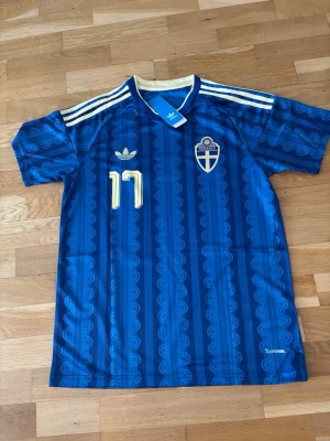  Sverige bortställ 2026 - Gyökeres #17 - Säljer en blå Sverige fotbollströja från Adidas med guldiga detaljer och nummer 17. Tröjan har klassiska vita Adidas-ränder på axlarna, broderad landslagssköld och namn Gyökeres på ryggen. Tillverkad i lätt och ventilerande material, perfekt för match eller träning.