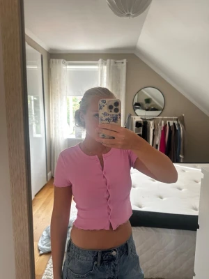 Rosa croppad topp med knappar - Söt rosa croppad topp med korta ärmar och knappar framtill. Toppen har en ribbad struktur och vågig kant nedtill som ger en trendig look. Perfekt att matcha med jeans eller kjol för en avslappnad stil.