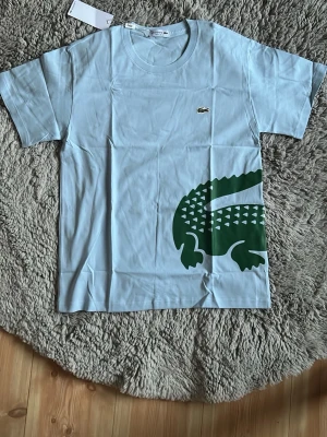 Lacoste t-shirt babyblue - En babyblå tröja med en krokodil som går genom magen, perfekt för att sticka ut under sommaren.  Bara skriva om de finns frågor!