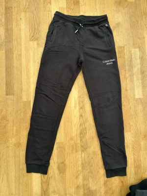 Svarta mjukisbyxor Calvin Klein Jeans - Svarta mjukisbyxor från Calvin Klein Jeans med broderad logga på vänster lår, dragsko i midjan och muddar vid benslut. Byxorna har två sidofickor och en bakficka. Perfekta för en chill och avslappnad stil.