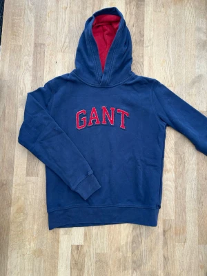 Marinblå hoodie från GANT - Snygg marinblå hoodie från GANT med röd logga broderad på bröstet. Insidan av huvan är röd vilket ger en cool kontrast. Hoodien har långa ärmar och ribbade muddar vid ärmslut och nederkant. Perfekt för en avslappnad och stilren look.
