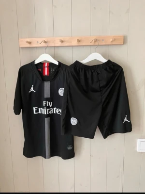 Svart PSG fotbollskit från Jordan - Snyggt svart fotbollskit från Jordan x PSG med tröja och shorts. Tröjan har Fly Emirates-tryck, PSG-logga och Jordan-logga. Shortsen har elastisk midja, PSG-märke och Jordan-logga. Perfekt för fotbollsträning eller match.