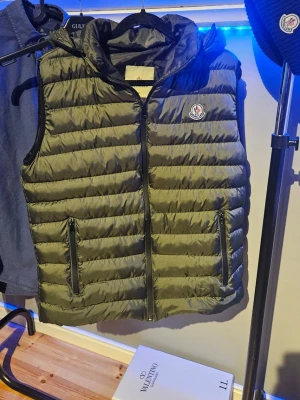 Grön dunväst från Moncler - Snygg olivgrön dunväst från Moncler med huva (Går att ta av) och dragkedja framtill. Västen har två praktiska fickor med dragkedja och det klassiska Moncler-logotypen på bröstet. Tillverkad i lätt, quiltat material som håller värmen utan att kännas tung.