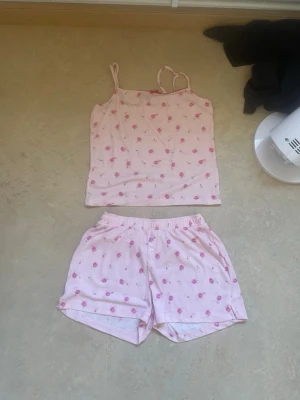 Rosa pyjamas-set med blommor - Sött pyjamas-set i ljusrosa med små rosa blommor. Setet består av ett linne med smala axelband samt både shorts och långa byxor med resår i midjan. Perfekt för mysiga kvällar hemma. Materialet känns mjukt och bekvämt mot huden. Oanvänd 