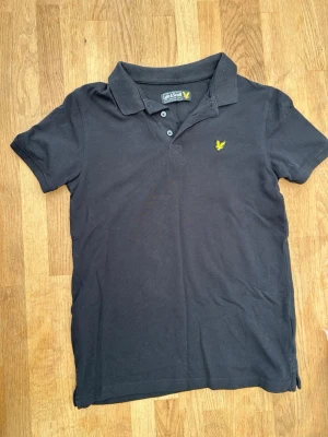 Svart pikétröja från Lyle & Scott - Klassisk svart pikétröja från Lyle & Scott med gul logga på bröstet. Tröjan har krage, knappar framtill och korta ärmar. Tillverkad i mjuk bomull för en bekväm känsla. Perfekt till jeans eller shorts.