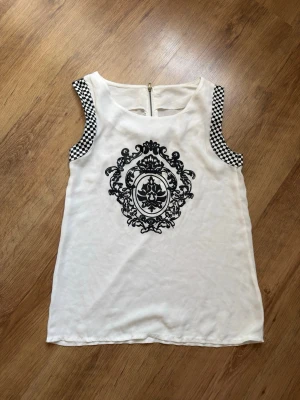 Vit ärmlös topp med svart tryck - Snygg vit ärmlös topp med svart barockinspirerat tryck framtill och schackrutiga detaljer vid ärmöppningarna. Toppen har rund halsringning och dragkedja bak. Perfekt för dig som vill sticka ut med coola detaljer.