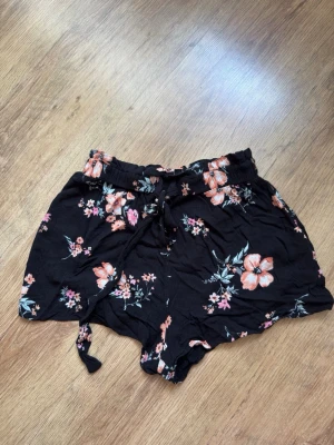 Blommiga shorts från H&M Divided - Svarta shorts från H&M Divided med blommigt mönster i rosa och orange toner. Modellen har hög midja, resår och snörning i midjan för extra komfort. Perfekta för varma dagar och en avslappnad stil.