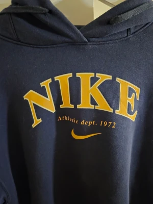 Mörkblå hoodie från Nike - Mörkblå hoodie från Nike med stor gul logga och texten 'Athletic dept. 1972' på bröstet. Klassisk huva med dragsko och ribbade muddar vid ärmar och nederkant. Perfekt för en sportig och avslappnad stil.