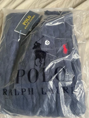 Marinblå kabelstickad tröja Polo Ralph Lauren - Kabelstickad marinblå tröja från Polo Ralph Lauren med rund halsringning och ribbade muddar. Klassisk röd broderad logga på bröstet. Perfekt för dig som gillar stilrena och tidlösa plagg. Materialet känns mjukt och håller värmen.