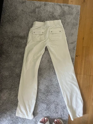 Vita mjukisbyxor från Juicy Couture - Säljer ett par vita mjukisbyxor från Juicy Couture i storlek XXS. Byxorna har raka ben, två fickor med lock bak och broderad JC-logga fram. Mjukt material som känns skönt mot huden och perfekt för en avslappnad stil.