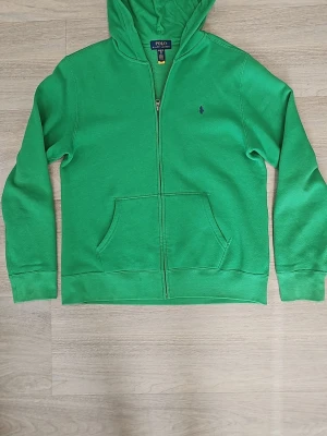 Grön hoodie från Polo Ralph Lauren - Snygg grön hoodie från Polo Ralph Lauren med dragkedja framtill och klassisk huva. Tröjan har känguruficka och den ikoniska lilla broderade loggan på bröstet. Tillverkad i mjuk bomullsblandning som känns skön mot huden. Perfekt för en avslappnad och stilren look. Storlek är XL men jag använde den en gång nör jag var 13 år. Pris kan diskuteras, hör gärna av er!