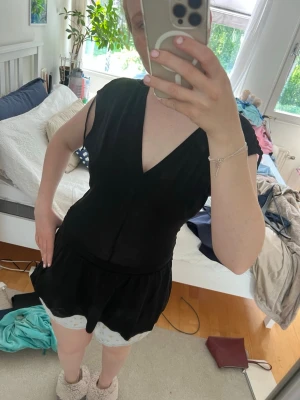 Svart topp med v-ringning - Snygg svart topp med v-ringning. Modellen har markerad midja och löst sittande shortsdel. Perfekt för en trendig och avslappnad look. Materialet ser ut att vara mjukt och lätt, vilket gör den bekväm att bära.
