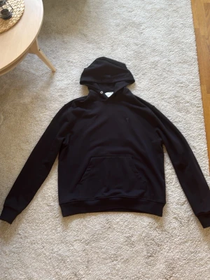  Ami Paris Hoodie - Svart hoodie från Ami Paris med klassisk känguruficka och broderad Ami de Coeur-logo på bröstet. Tillverkad i mjuk bomull, har huva med dragsko och ribbade muddar vid ärmar och nederkant. Perfekt för en clean och stilren look. Använd sällan och är i bra skick. Tagit ut snören då de va långa. Pris kan diskuteras.