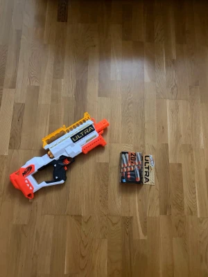 Nerf Ultra leksaksgevär med pilar - Säljer ett Nerf Ultra leksaksgevär i vitt, orange och svart med tillhörande magasin och extra pilar. Geväret har en cool design med detaljer i gult och är perfekt för roliga Nerf-strider. Paketet inkluderar även en förpackning med 20 Ultra-pilar.