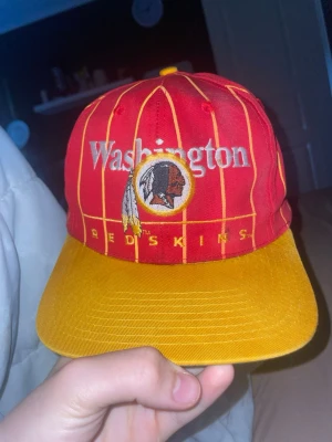 Washington Redskins röd/gul keps - Säljer en retro snapback-keps med Washington Redskins broderat framtill. Kepsen är röd med gula ränder och har en gul skärm. Klassisk logga och text på framsidan, grön undersida på skärmen och MLB-märke på insidan. Perfekt för dig som gillar sportig vintage-stil.