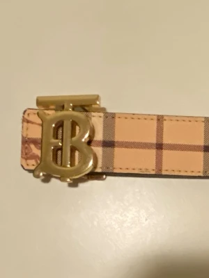 Burberry vändbart bälte med guldspänne - Säljer ett vändbart Burberry-bälte med klassiskt rutigt mönster på ena sidan i beige och brunt, och svart rutigt mönster på andra sidan. Bältet har ett stort guldfärgat TB-spänne och är gjort i skinn. Perfekt accessoar för att lyfta din outfit.