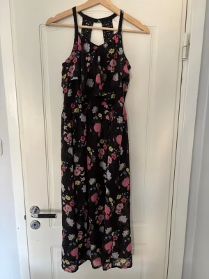 Svart blommig långklänning med volang - Storlek 146 man är större i passformen.            Svart långklänning med blommigt mönster i rosa, vitt och gult. Klänningen har smala axelband, volangdetalj framtill och en snygg öppen rygg med dekorativa band. Perfekt för sommarens alla tillfällen.