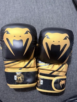 Svarta och guldiga boxningshandskar Venum - Säljer ett par snygga boxningshandskar från Venum i svart och guld. Handskarna har ett stort ormmotiv på ovansidan och bred kardborrestängning för extra stöd. Tillverkade i slitstarkt syntetmaterial, perfekta för träning och sparring.