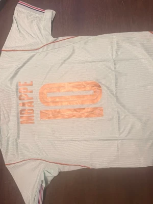 Ljusblå Nike fotbollströja Mbappé #10 - Snygg ljusblå fotbollströja från Nike med guldiga detaljer och tryck. Tröjan har Mbappé och nummer 10 på ryggen, samt Frankrikes landslagsemblem på bröstet. Korta ärmar med randiga muddar och guld Nike-logga framtill. Perfekt för match eller träning.