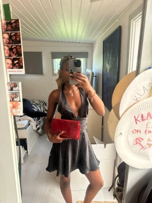 Svart halterneck kort klänning - Snygg svart kort klänning med halterneck och djup v-ringning. Klänningen har en lätt utsvängd kjol och slät, glansig yta som ger en elegant look. Perfekt för dig som vill sticka ut med en stilren och trendig outfit.