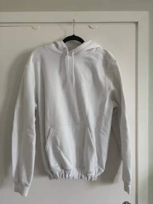 Vit basic hoodie med huva - En clean vit hoodie med huva och dragsko, samt en stor känguruficka framtill. Hoodien har ribbade muddar vid ärmar och midja för en skön passform. Perfekt för en avslappnad och stilren look. Hoodien har ett resårband nertill men det går att ta bort om man vill.