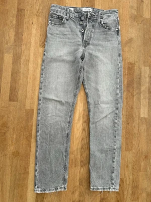 Grå raka jeans från Jack & Jones - Säljer ett par grå raka jeans från Jack & Jones med klassisk femficksmodell och knäppning fram. Jeansen har råa, fransiga benslut och en ljus tvätt som ger en avslappnad vibe. Tillverkade i bomull för skön känsla och snygg passform. W27 L32