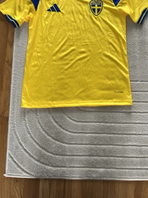 Svensk landslagströja Adidas gul - Gul svensk landslagströja från Adidas med blå detaljer och det svenska fotbollsförbundets emblem på bröstet. Tröjan har korta ärmar och är tillverkad i ett lätt och ventilerande material, perfekt för träning eller match.