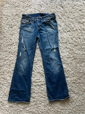 Blå bootcut Dieseljeans med slitningar - Säljer ett par blå bootcut Dieseljeans med slitna detaljer och råa kanter. Jeansen har dekorativa sömmar och nitar på bakfickorna samt klassisk femficksmodell. Perfekta för dig som gillar en avslappnad och trendig stil.