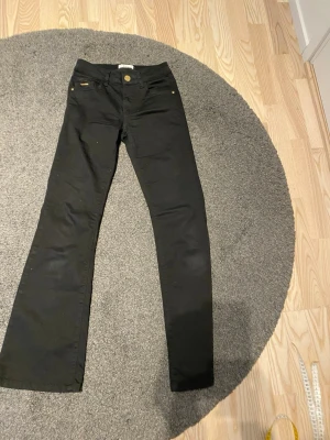 Svart  Lido Flare  jeans från Isay st 32 - Snygg svart byxa med svagt utsvängda ben nedtill. Läderjapp bag med loggan Isay snygg detalj med pärlor på ena framfickan. Metallknapp och dragkedja. Fyra fickor. 68 cm i midjemått och beninnerlängd  75 cm. Skickas nytvättad.