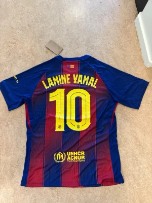 Barcelona matchtröja Lamine Yamal #10 - Säljer en officiell FC Barcelona matchtröja med Lamine Yamal och nummer 10 på ryggen. Tröjan har klassiska blå och vinröda ränder, klubbmärke på bröstet och UNHCR-logga. Tillverkad i lätt och ventilerande Dri-FIT-material, perfekt för fotboll eller träning.