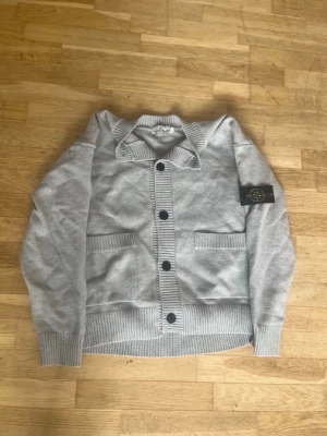 Grå kofta från Stone Island - Snygg grå kofta från Stone Island med ribbstickade muddar, stora knappar och två framfickor. Klassisk logotyp på ärmen och skön krage. Perfekt för lager-på-lager och streetwear-stil.