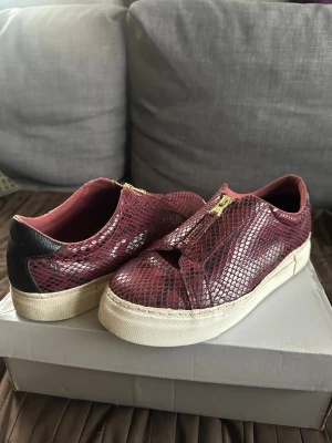 Vinröda sneakers med ormmönster - Snygga vinröda sneakers med ormmönstrad yta och guldfärgade dragkedjor framtill. Skorna har vit sula och svart detalj på hälen. Materialet ser ut som konstläder med reptilstruktur för en cool och unik look.