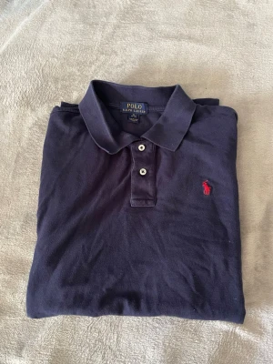 Mörkblå pikétröja från Ralph Lauren - Klassisk mörkblå pikétröja från Ralph Lauren med krage, två knappar och den ikoniska röda logotypen broderad på bröstet. Tillverkad i mjuk bomull som känns skön mot huden. Perfekt till jeans eller shorts för en clean look.