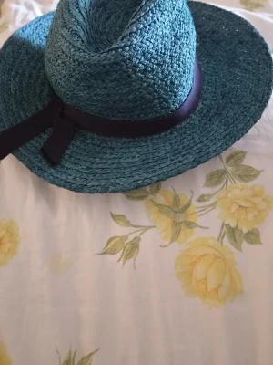Blågrön stråhatt med svart band - Snygg blågrön stråhatt från Inwear med brett brätte och svart band runt kronan. Hatten har en klassisk form och är tillverkad i flätat stråmaterial. Perfekt för soliga dagar och ger en cool touch till din outfit.